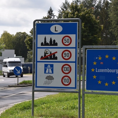 Das Grenzschild von Luxemburg, eingefasst von Europa-Sternen auf blauem Grund, aufgenommen an der deutsch-luxemburgischen Grenze neben einem Schild mit Geschwindigkeitsbegrenzungen.