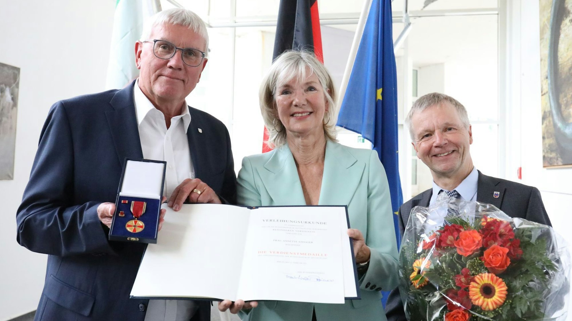 Drei Männer und eine Frau halten den Orden, eine Urkunde und einen Blumenstrauß in den Händen.