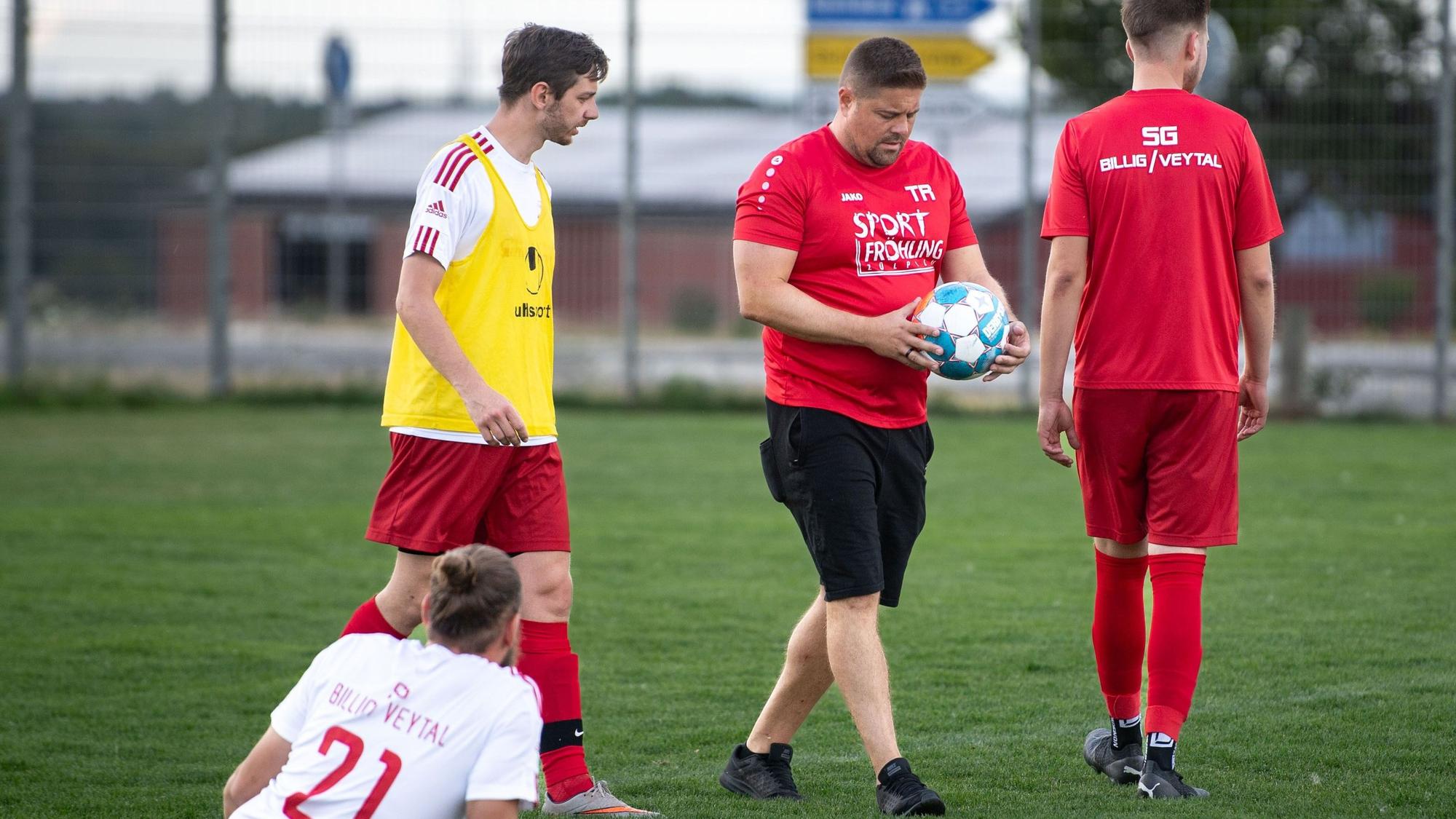 Das Bild zeigt einen Fußballtrainer, der einen Ball in den Händen hält. Zwei Spieler stehen neben ihm, ein weiterer sitzt auf dem Rasen.