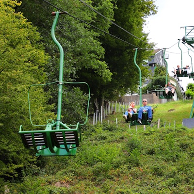 Menschen fahren in einem Sessellift einen begrünten Hang hinauf und hinunter.