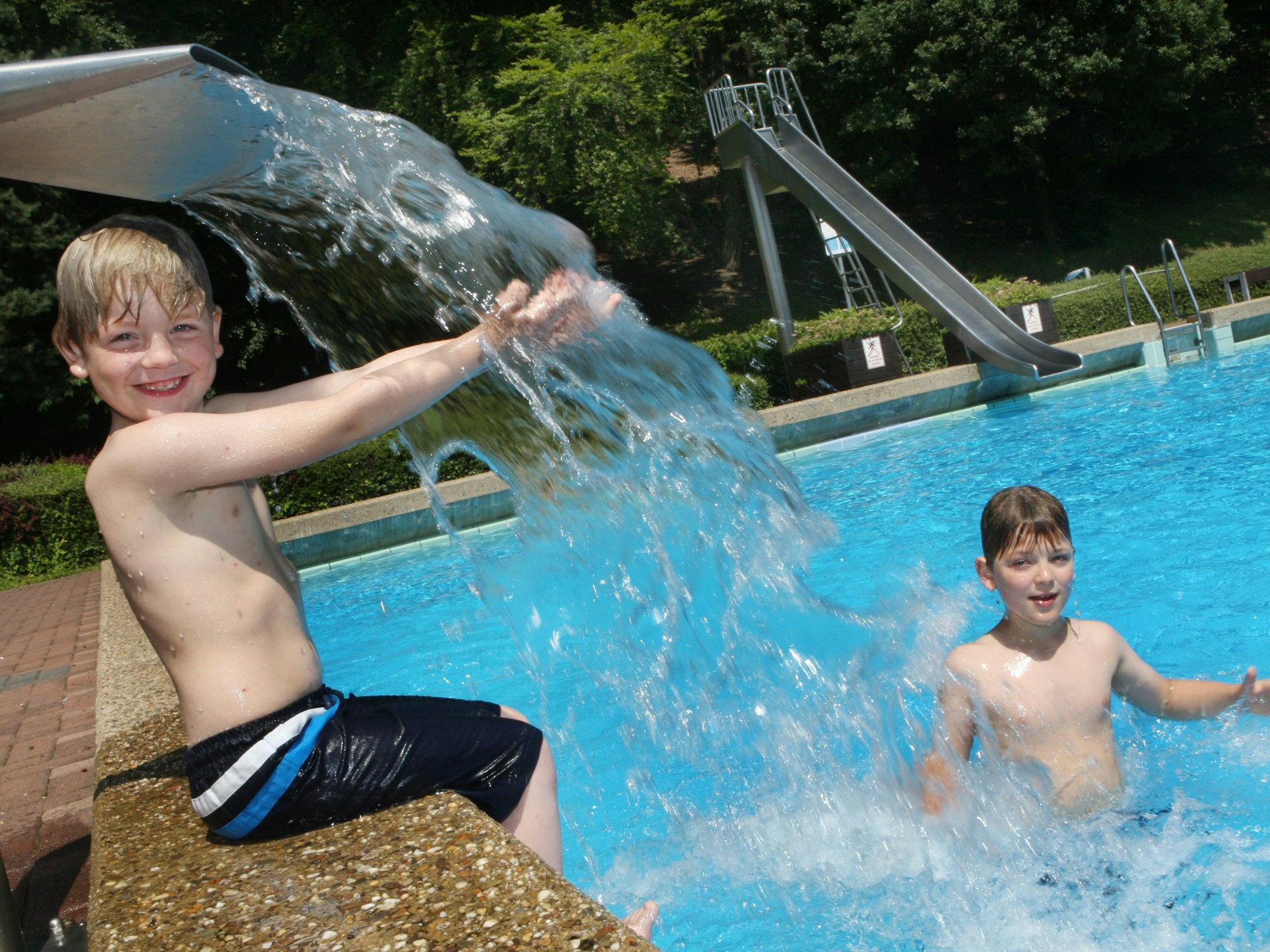 Zwei Kinder spielen an einem Wasserspeier in einem Freibad. Im Hintergrund ist eine Rutsche zu sehen.