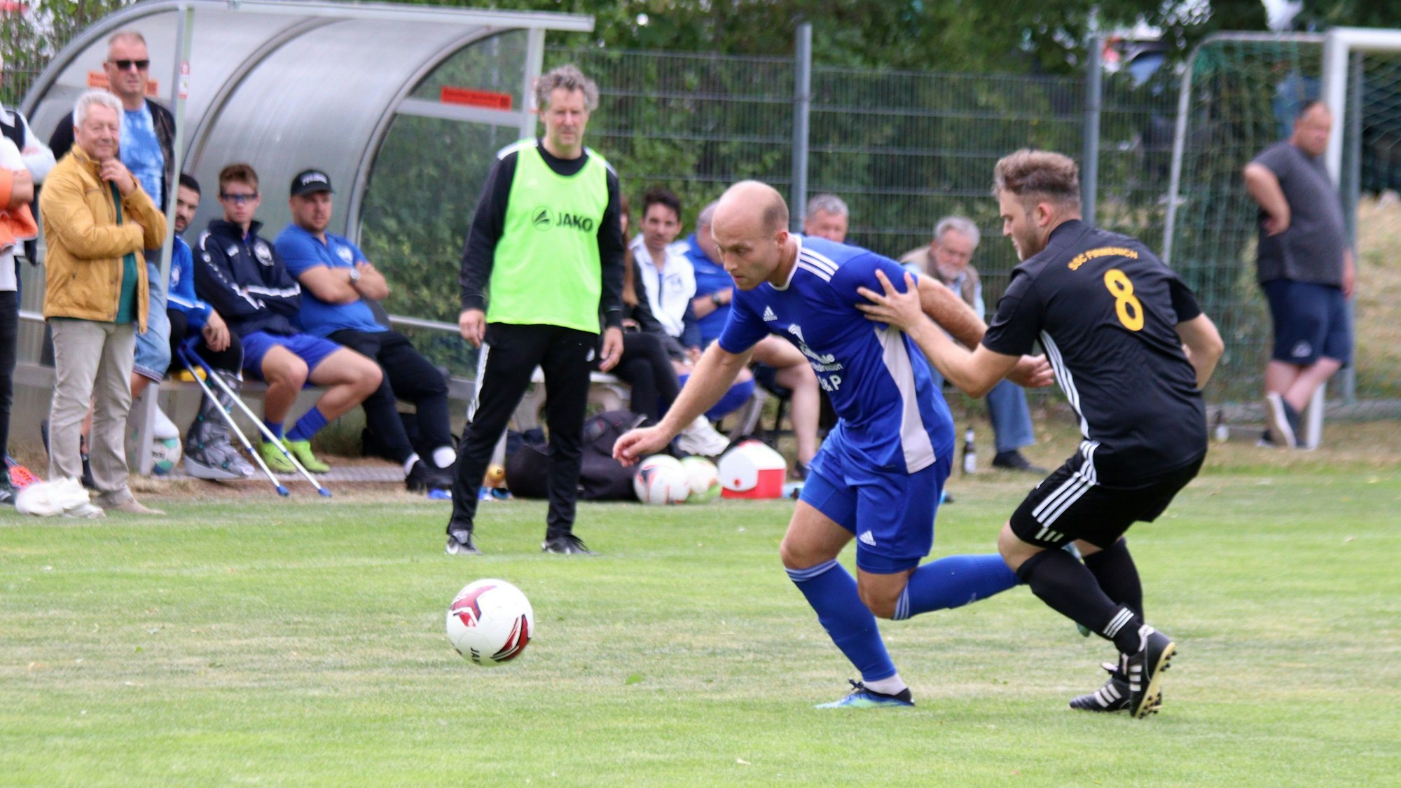 Weilerswists Felix Kortholt zieht an Luca Riedling vorbei. Im Hintergrund beobachten Trainer Horst Bartz im gelben Leibchen und die Zuschauer die Aktion.