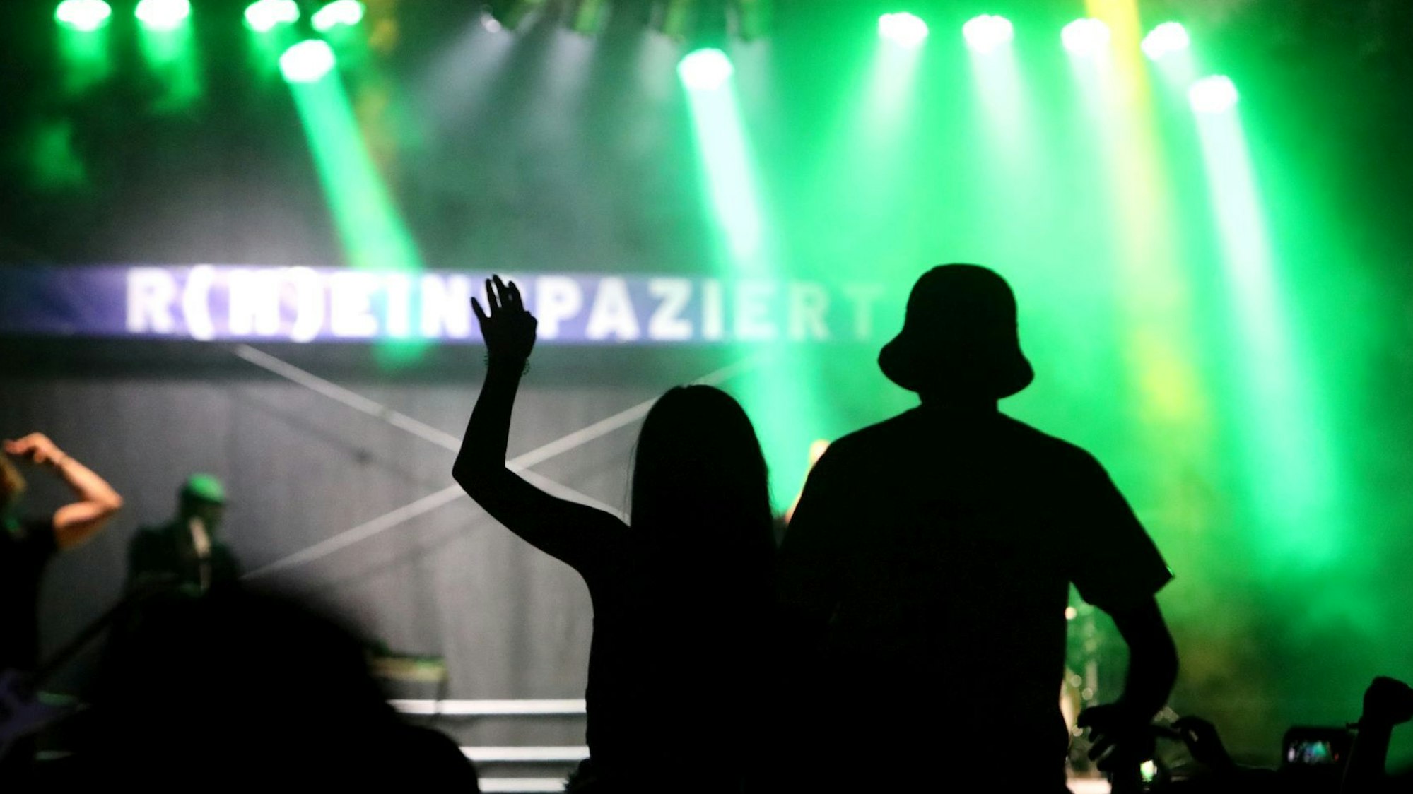 Der Schattenriss von Zuschauern vor der mit grünen Scheinwerfern beleuchteten Bühne mit dem Schriftzug „R(h)einspaziert“.
