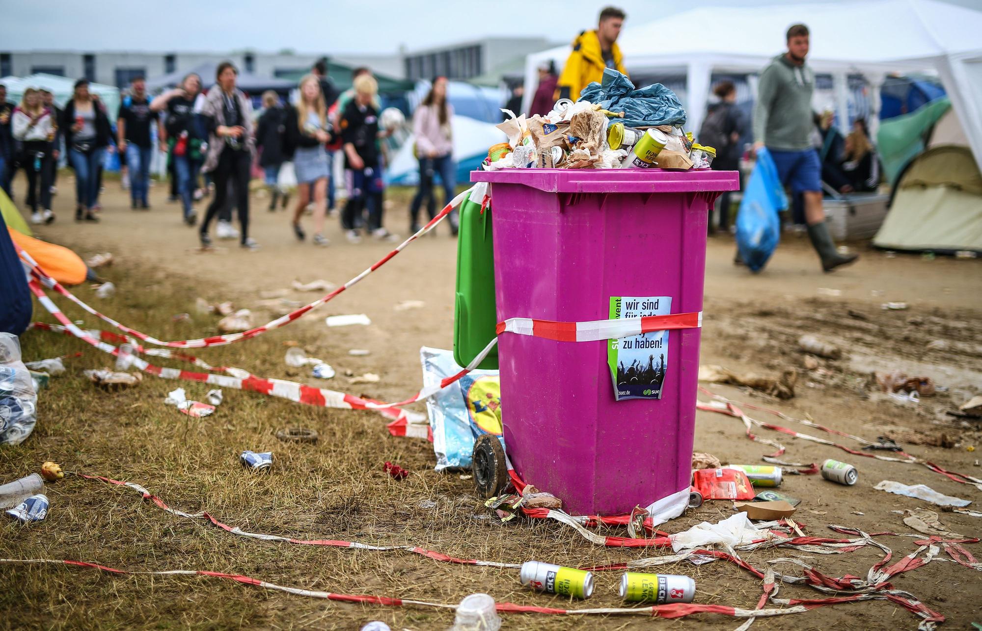 Blick auf eine Wiese, die voller Bierdosen, Absperrbänder und Verpackungsmüll ist Mittig eine überquellende pinkfarbene Mülltonne, im Hintergrund Menschen.