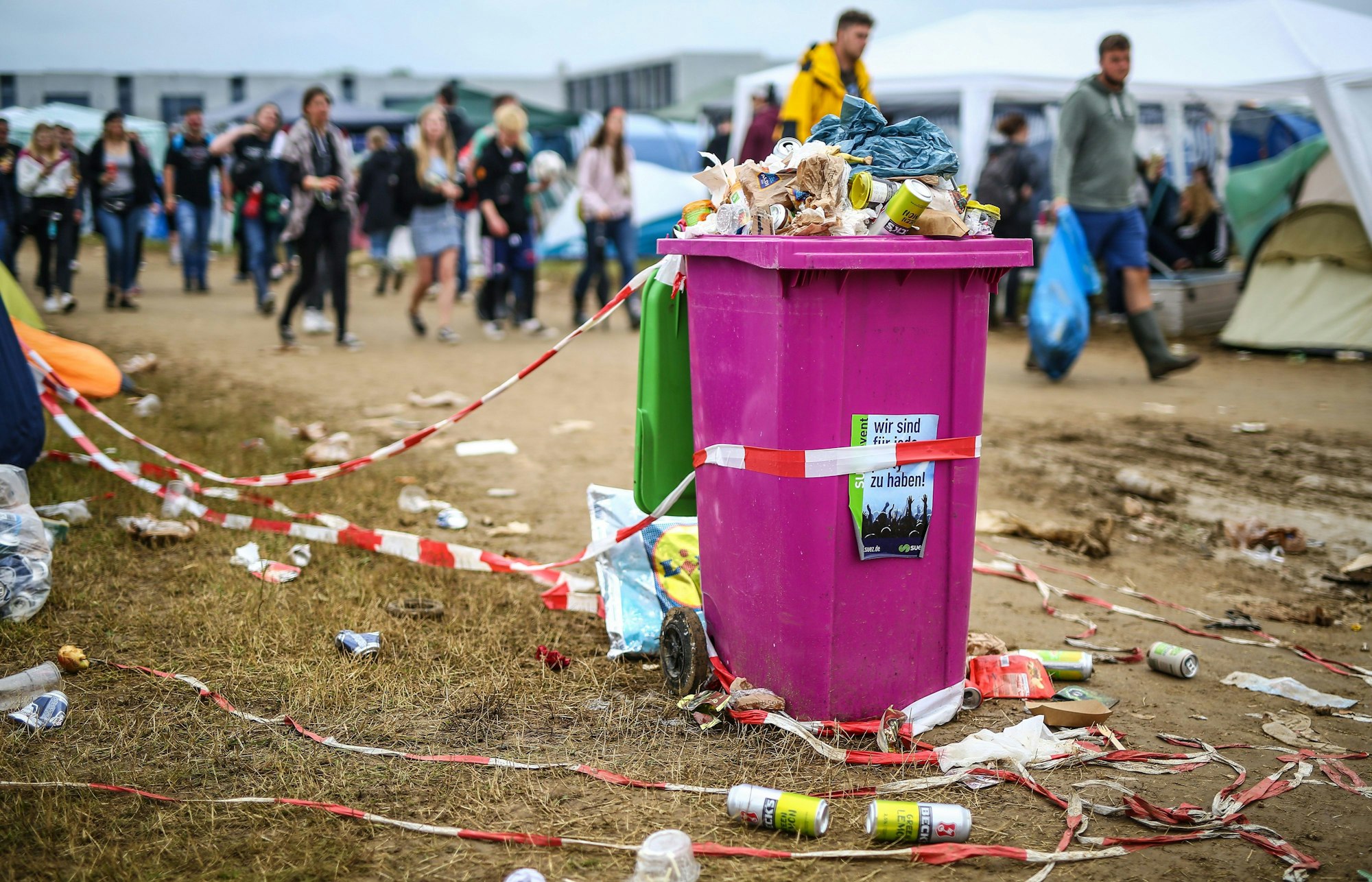 Blick auf eine Wiese, die voller Bierdosen, Absperrbänder und Verpackungsmüll ist Mittig eine überquellende pinkfarbene Mülltonne, im Hintergrund Menschen.