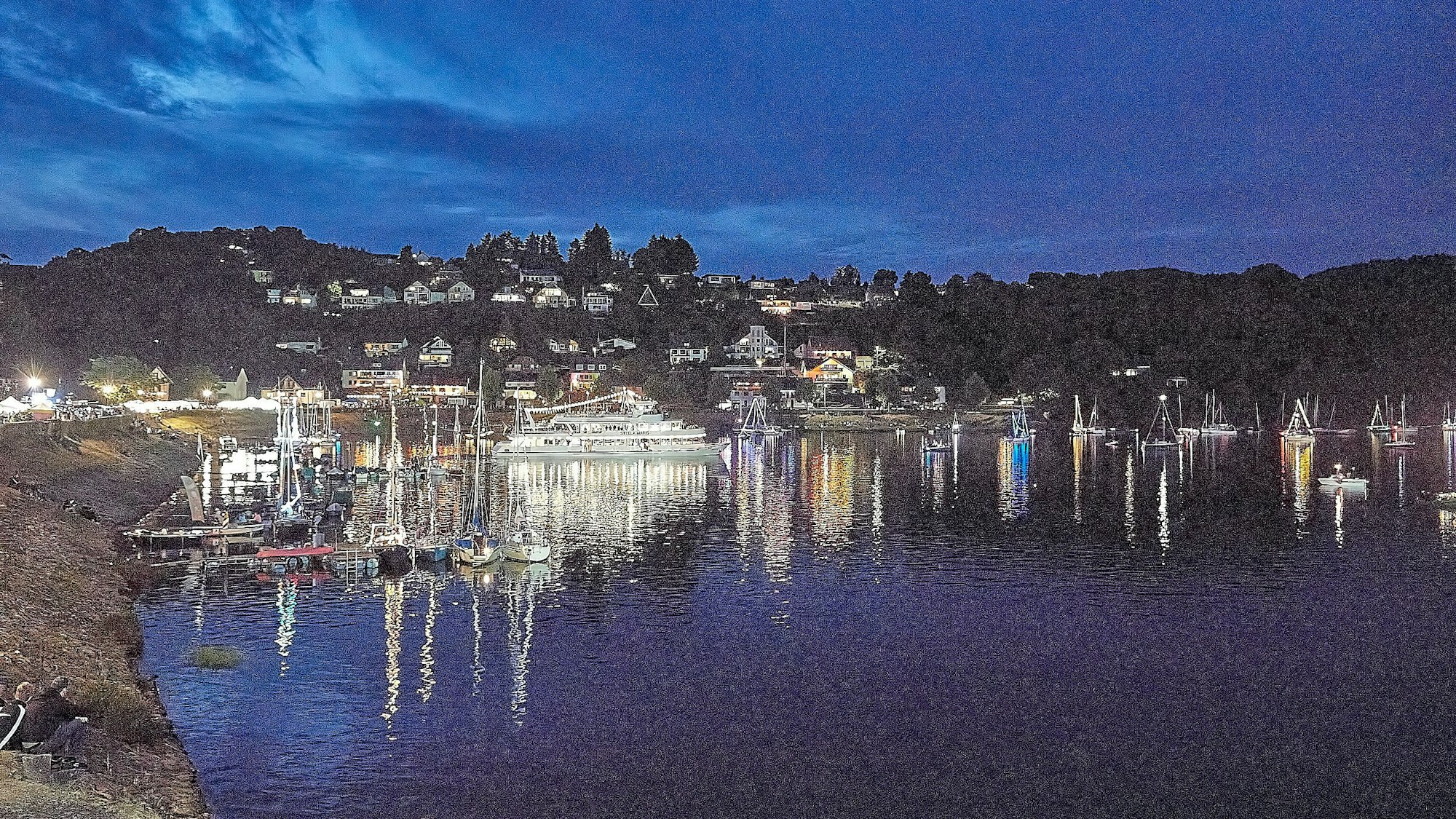Blick über die Bucht, in der zahlreiche erleuchtete Boote und Schiffe bei hereinbrechender Dunkelheit den Beginn des Feuerwerks erwarten.