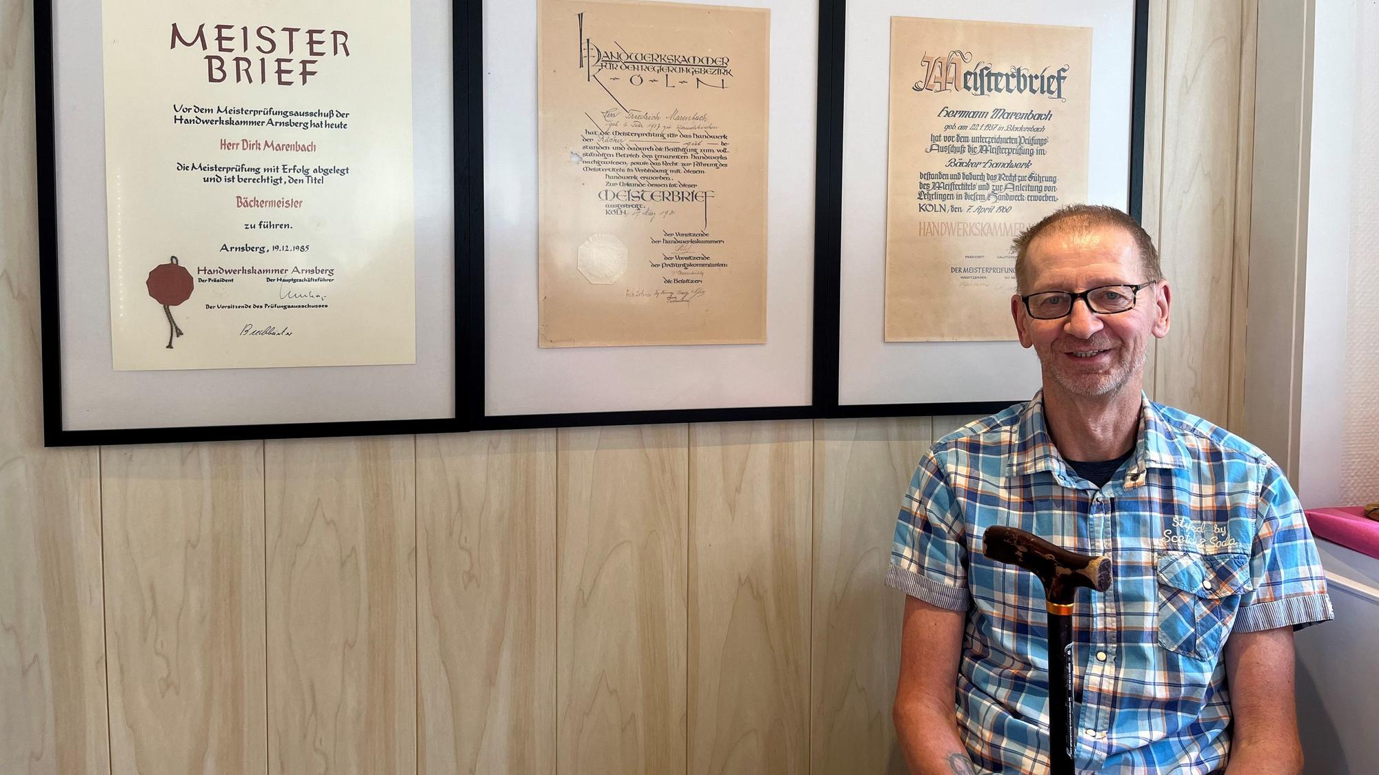 Ein Mann mit Gehstock sitzt vor mehreren Meisterbriefen, die an der Wand hängen.