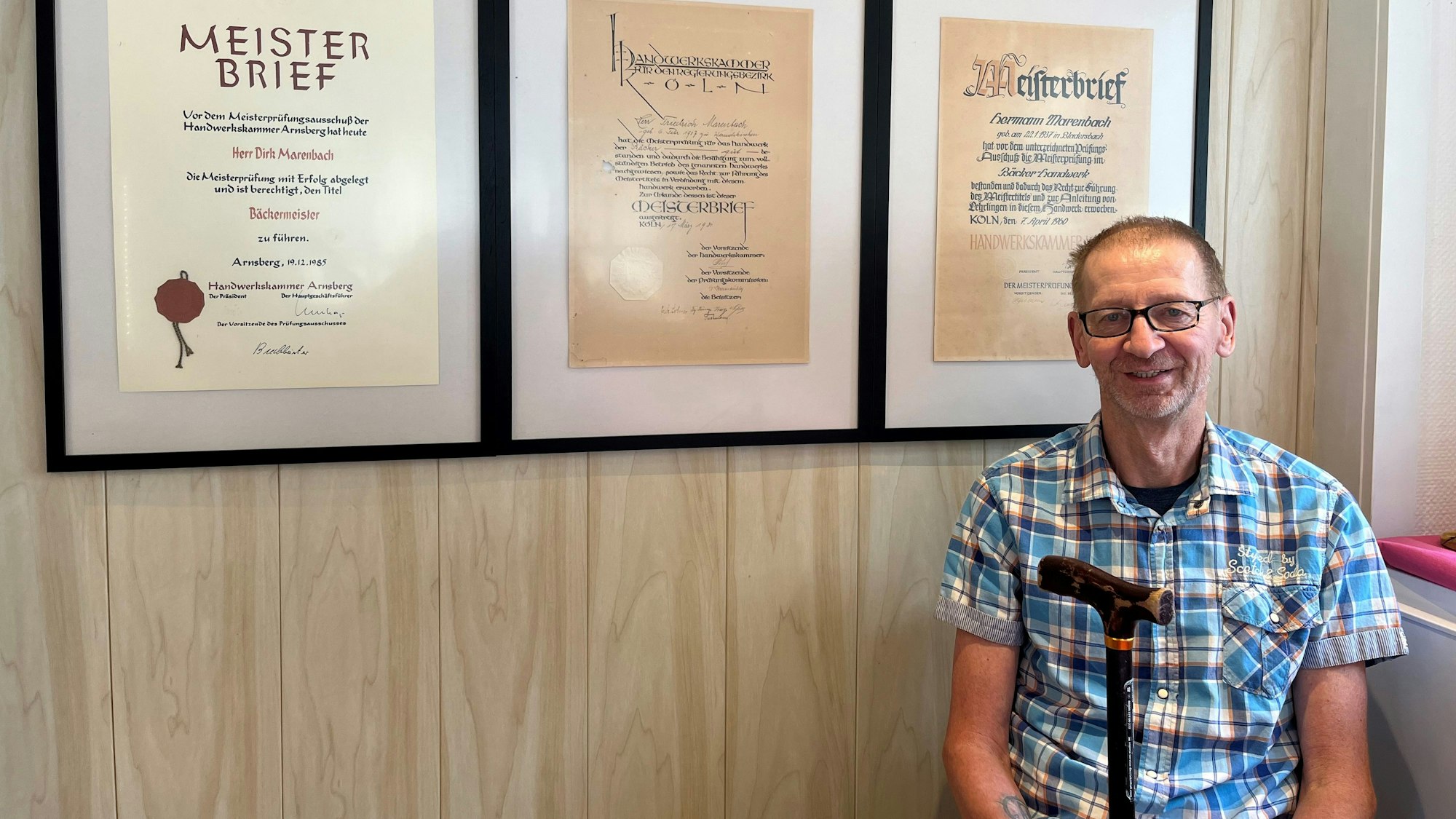 Ein Mann mit Gehstock sitzt vor mehreren Meisterbriefen, die an der Wand hängen.
