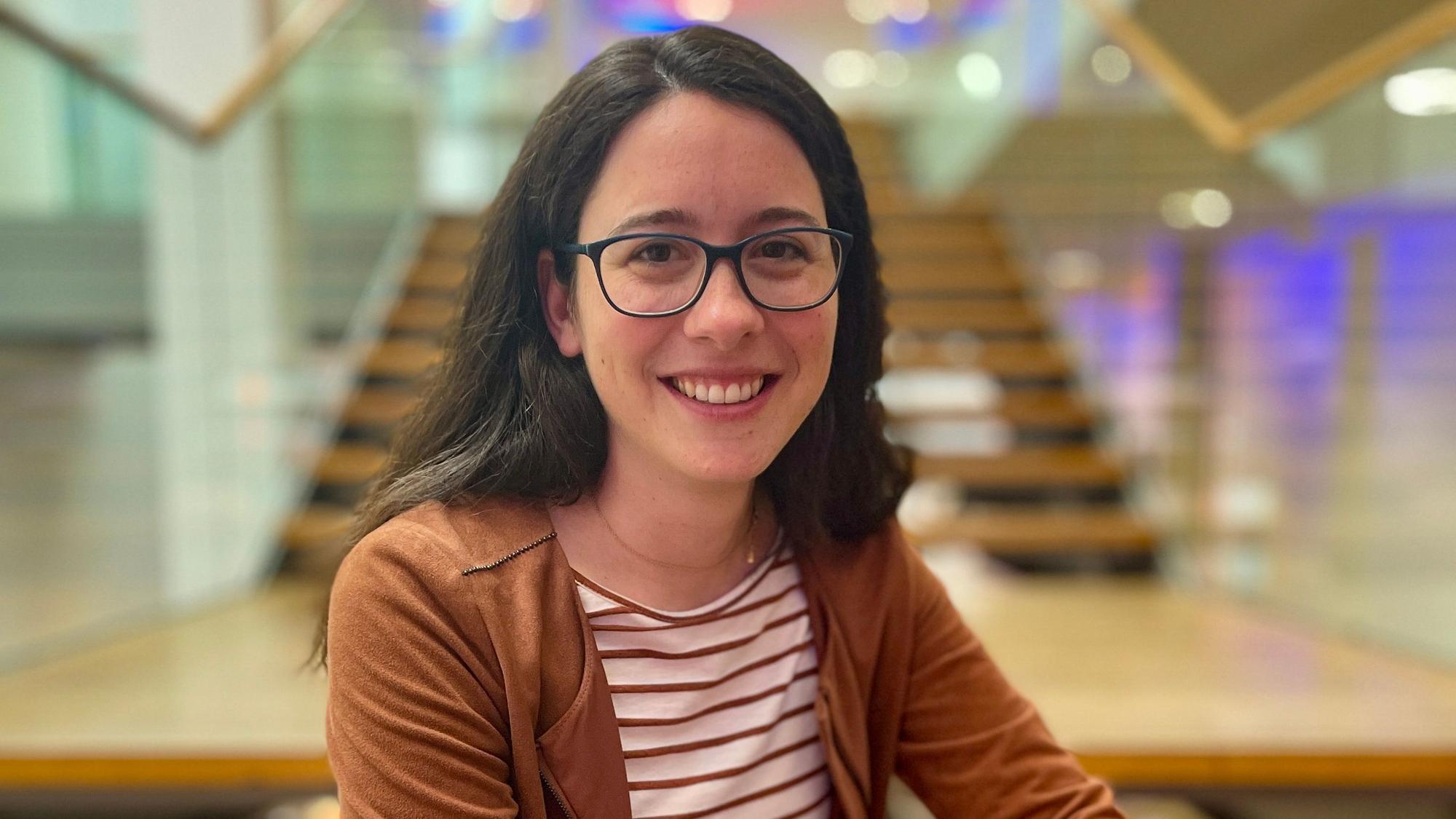 Auf dem Foto ist eine junge Frau mit dunklem Haar und einer schwarzen Brille zu sehen.