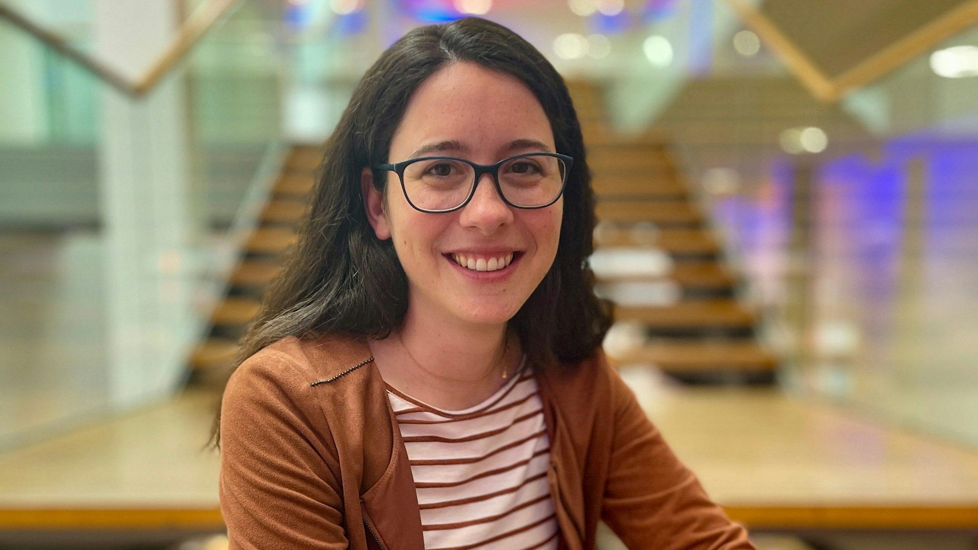 Auf dem Foto ist eine junge Frau mit dunklem Haar und einer schwarzen Brille zu sehen.