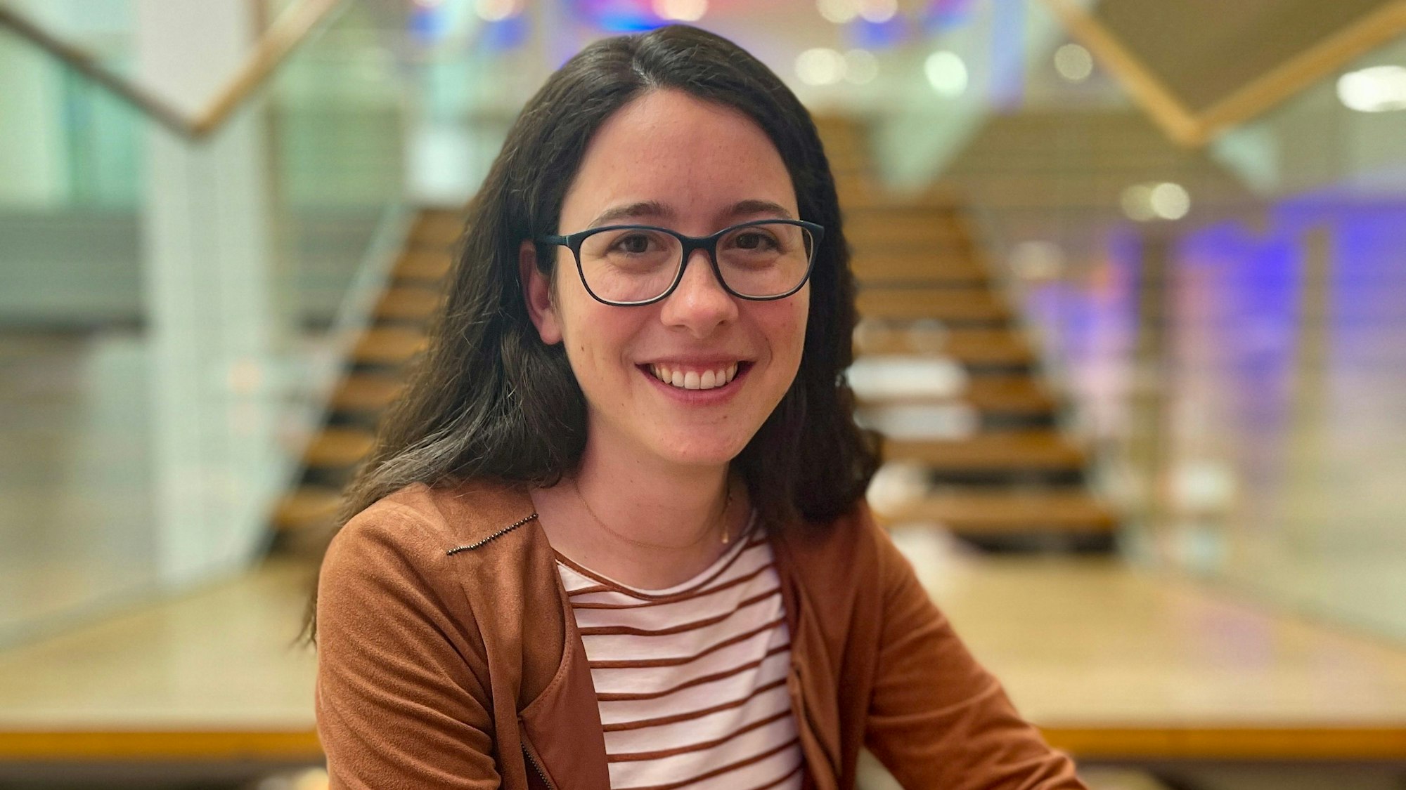 Auf dem Foto ist eine junge Frau mit dunklem Haar und einer schwarzen Brille zu sehen.