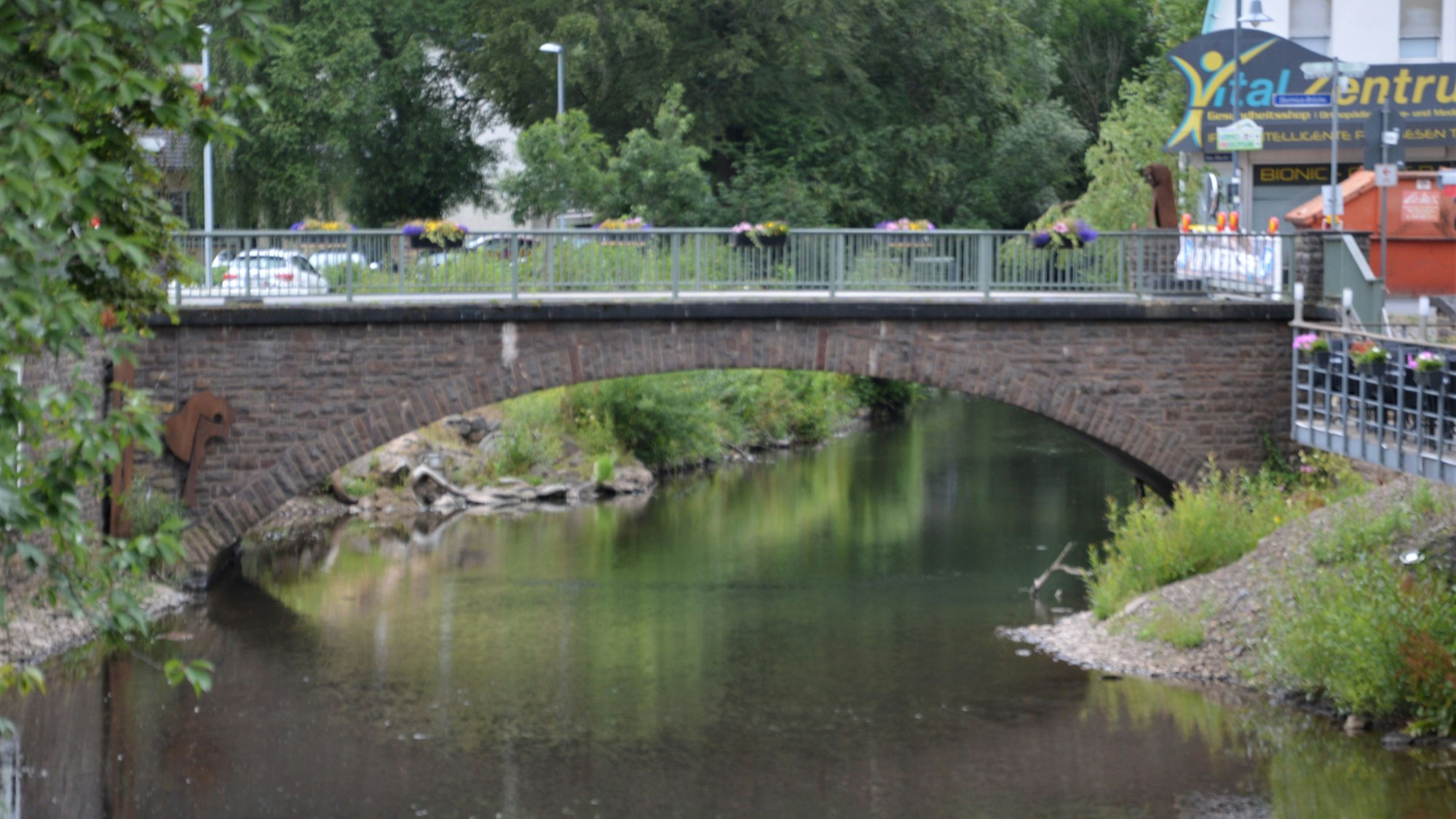 Zu sehen ist die beschädigte Brücke über die Olef in Schleiden. Am Geländer hängen Blumenkübel, links im Bild sind Bäume zu erkennen.