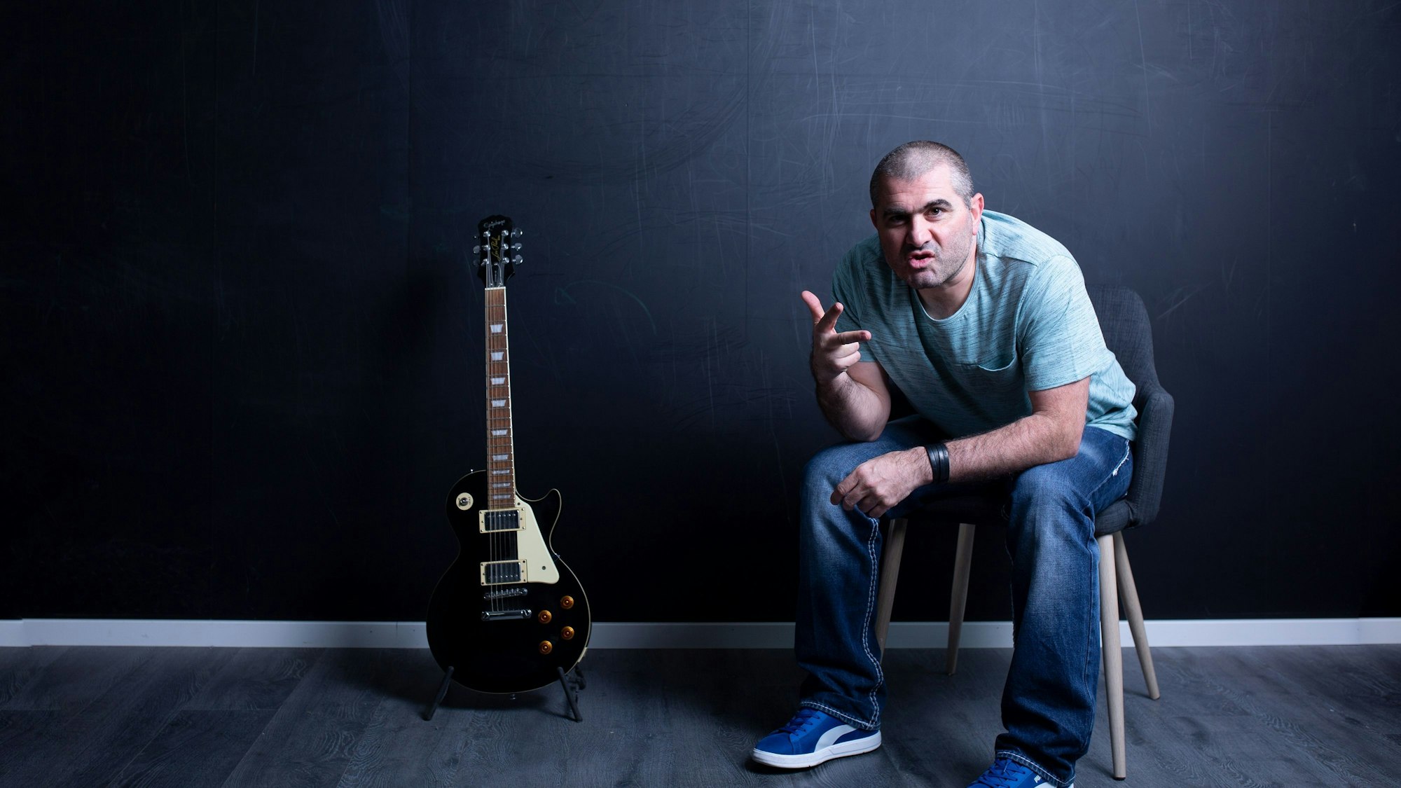 Ein Mann sitzt in einem leeren Raum auf einem Stuhl. Neben ihm steht eine E-Gitarre auf dem Boden.
