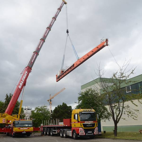 Ein großer Autokran hebt ein langes orange lackiertes Metallteile über einen Tieflader und eine Fabrikhalle hinweg.