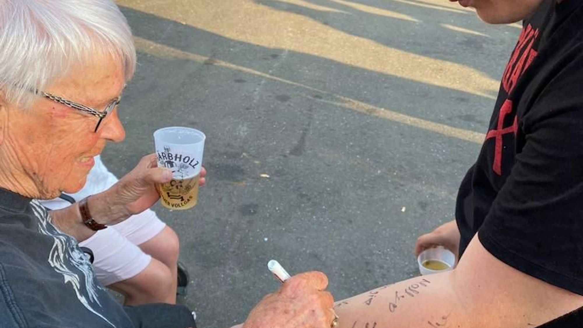Eine Frau unterschreibt auf dem Arm eines Jugendlichen. Sie hält ein Bier in der Hand.
