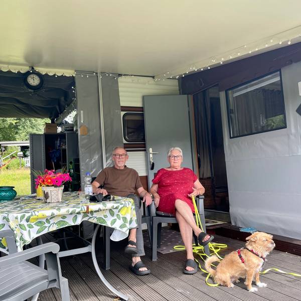 Zwei Senioren mit Hund sitzen in Campingstühlen vor ihrem Dauercamper-Wohnwagen.