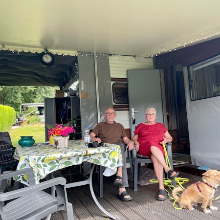 Zwei Senioren mit Hund sitzen in Campingstühlen vor ihrem Dauercamper-Wohnwagen.