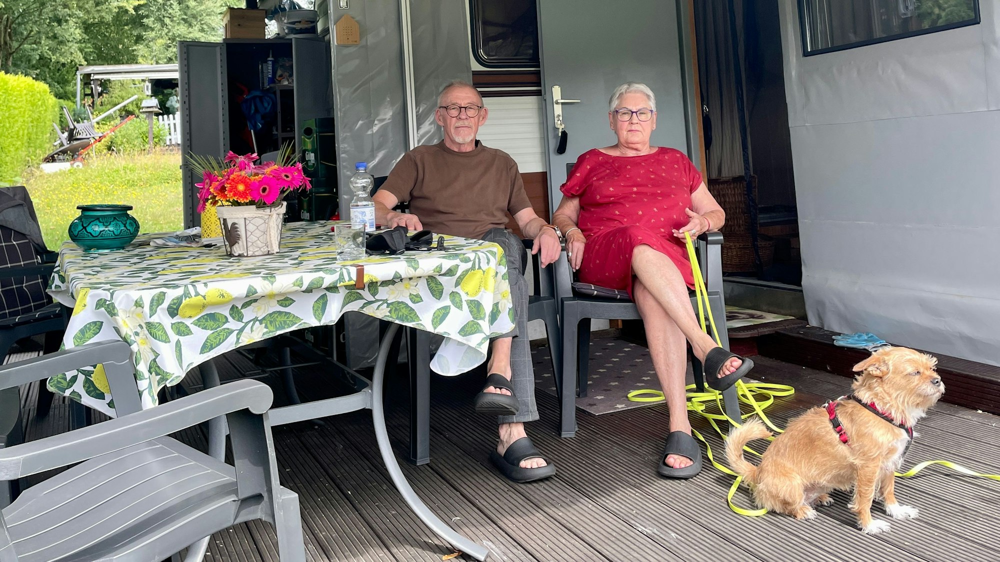 Zwei Senioren mit Hund sitzen in Campingstühlen vor ihrem Dauercamper-Wohnwagen.