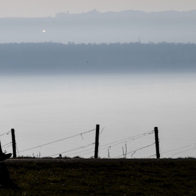 Jogger laufen auf einem Wanderweg in Richtung Meersburg, während im Hintergrund die Stadt Konstanz am Bodensee im Dunst zu sehen ist.