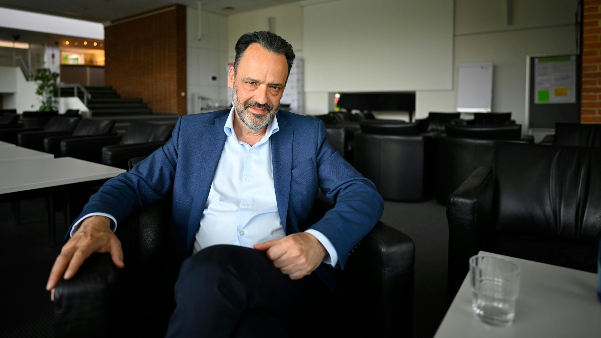 Ein Mann im Anzug sitzt in einem Konferenzraum auf einem Stuhl und schaut in die Kamera.