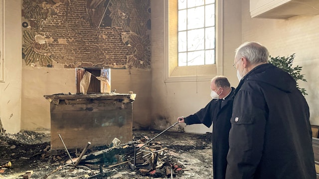 Zwei Priester stehen in einem Kircheninnenraum vor Überrresten nach einem Brand.