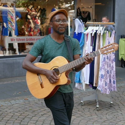 Ein junger Mann mit Schirmmütze und Gitarre; er trägt eine Brille und hat eine dunkle Hautfarbe.