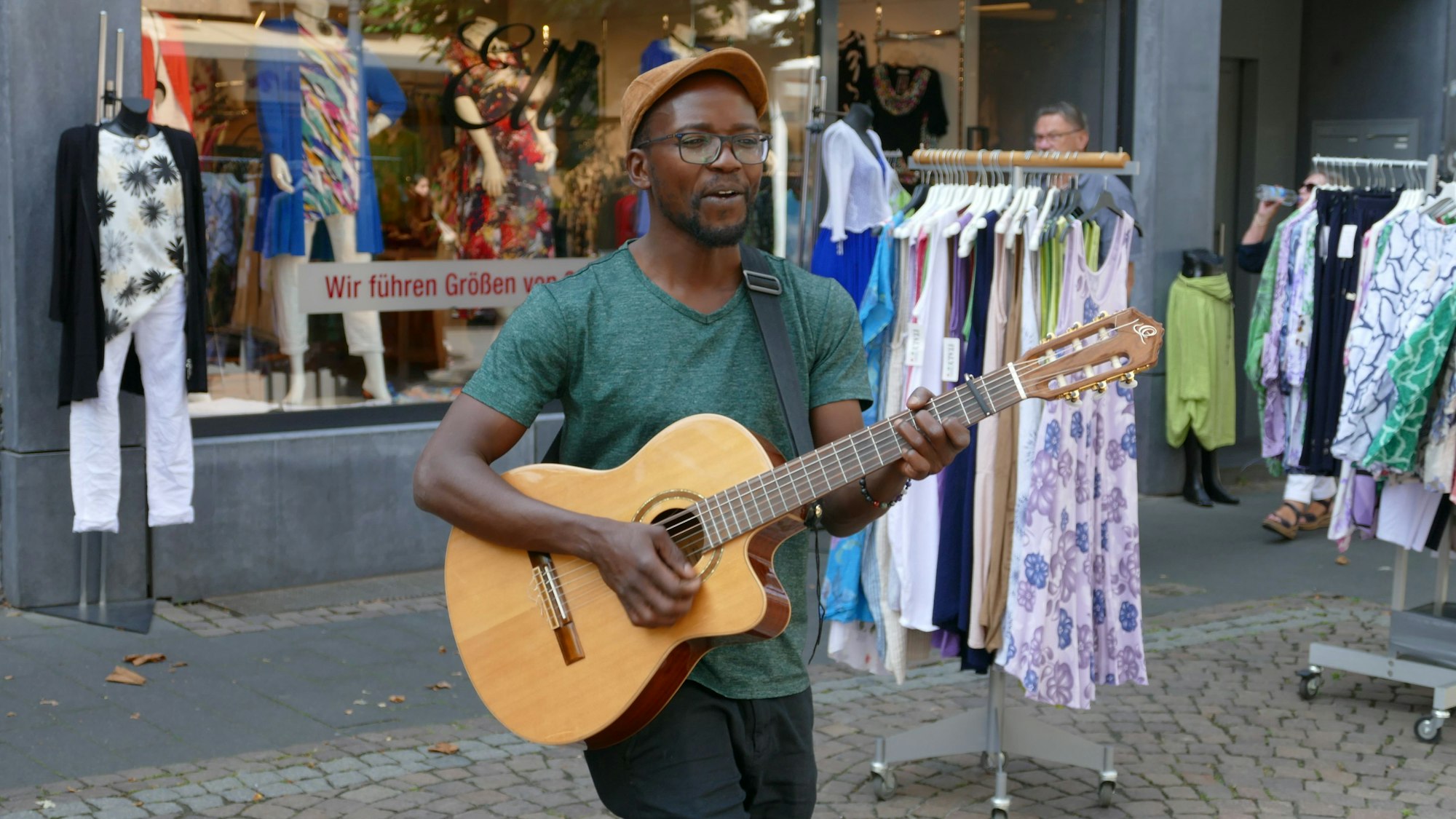 Ein junger Mann mit Schirmmütze und Gitarre; er trägt eine Brille und hat eine dunkle Hautfarbe.