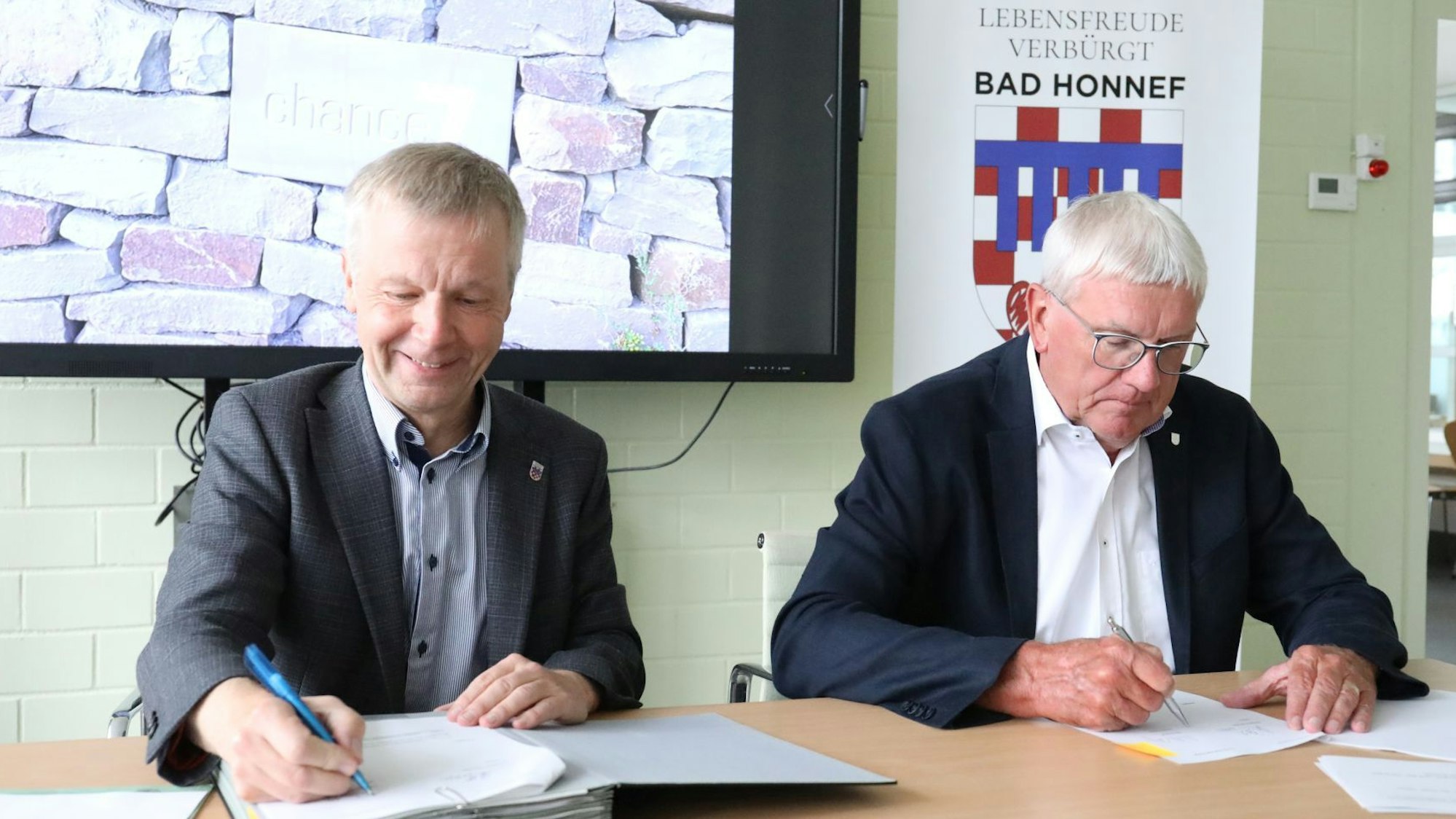 Zwei Männer sitzen nebeneinander an einem Tisch und unterschreiben die Verträge. Im Hintergrund das Stadtlogo mit dem Motto „Lebensfreude verbürgt“.