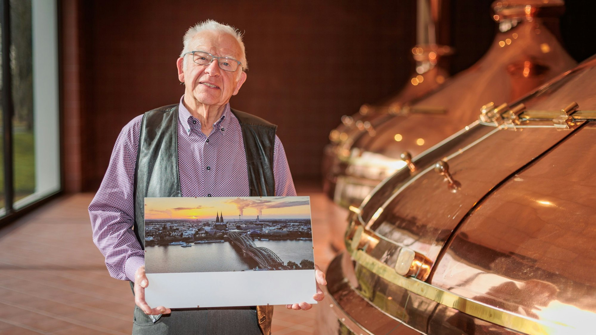 Ein älterer Mann hält ein Foto in den Händen, das den Kölner Dom, den Rhein und die Hohenzollernbrücke aus der Luftperspektive zeigt.