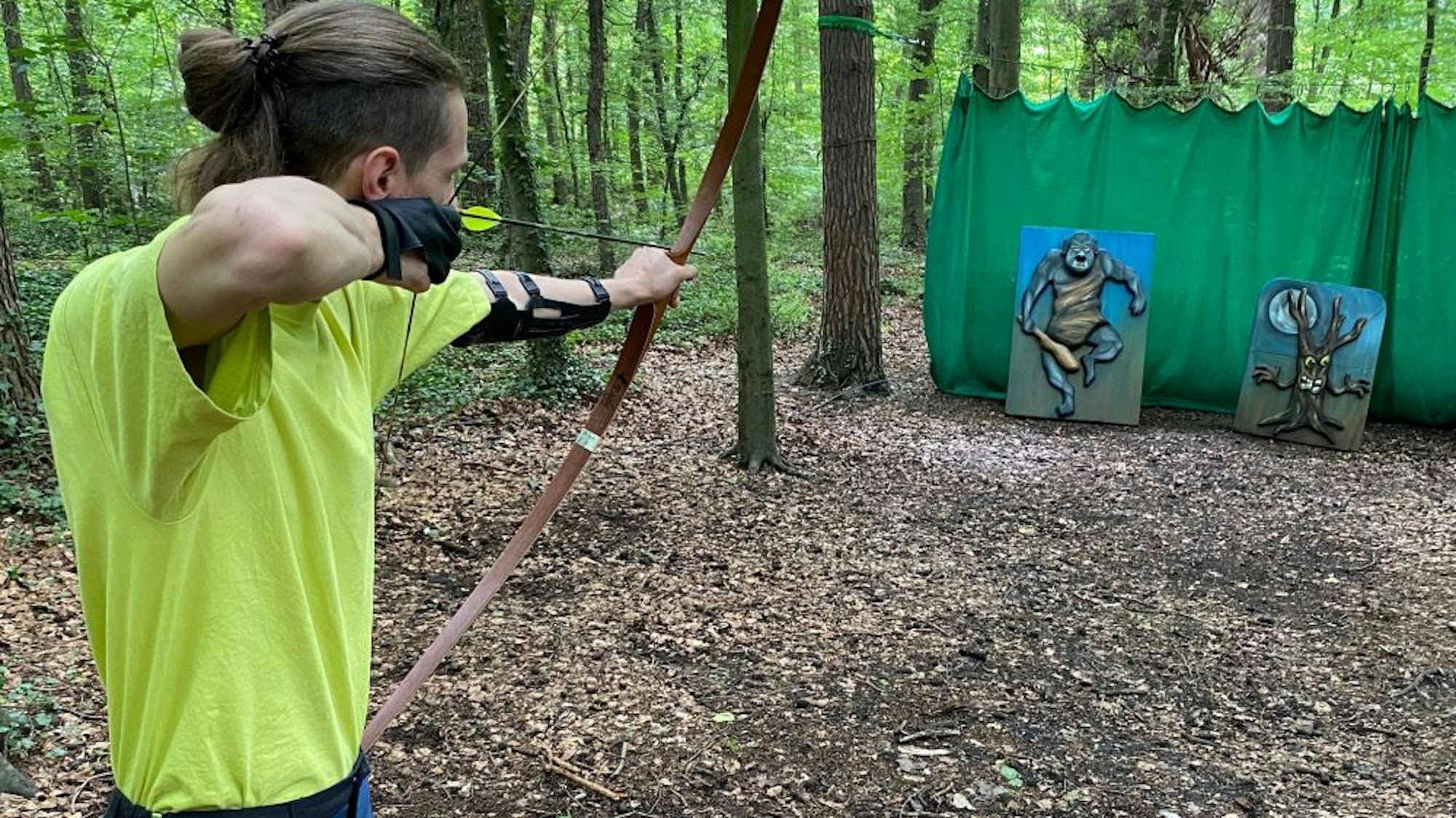 Ein junger Mann spannt den Bogen und zielt auf Scheiben, auf denen Fabelwesen zu sehen sind.