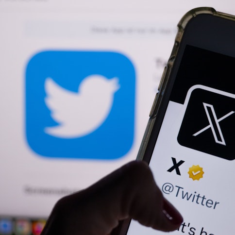 Auf dem offiziellen Profil von Twitter auf dem Bildschirm eines Smartphones ist der weiße Buchstabe X auf schwarzem Hintergrund zu sehen, während im Hintergrund das bisherige Logo des Kurznachrichten-Dienstes Twitter angezeigt wird.