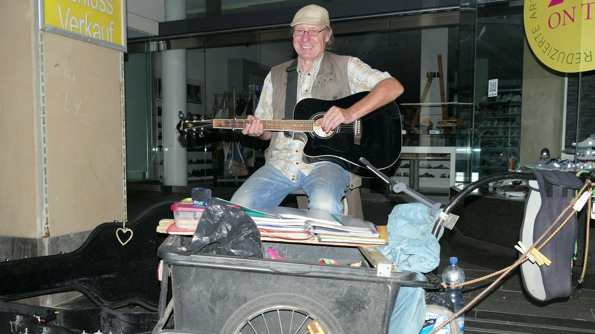 Ein Mann mit Schirmmütze und Gitarre, vor ihm steht ein Fahrradanhänger. Er sammelt Geld im aufgeklappten Gitarrenkoffer.