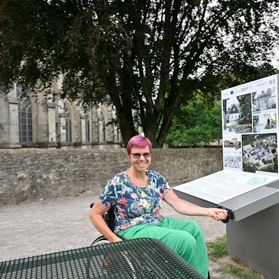 Eine Frau sitzt im Rollstuhl und bedient lachend eine Kurbel, mit der eine Infostation am Altenberger Dom bedient wird.