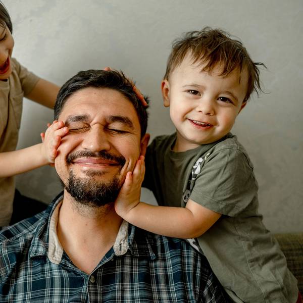 Ein Vater (mittig) sitzt mit seinen beiden Kindern (links und rechts) auf einem Sofa. Alle drei lächeln.
