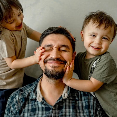 Ein Vater (mittig) sitzt mit seinen beiden Kindern (links und rechts) auf einem Sofa. Alle drei lächeln.