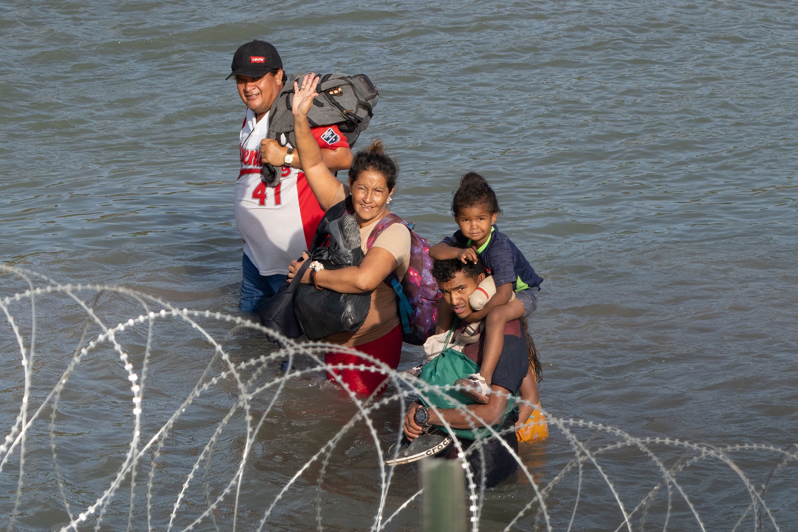 Migranten winken vor Stacheldraht als sie durch den Rio Grande laufen.