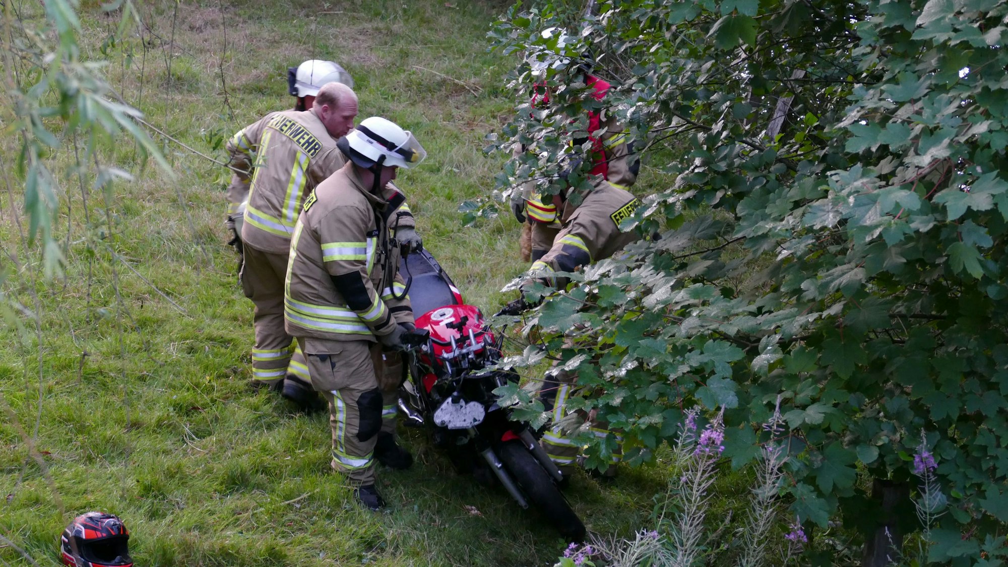 Einsatzkräfte der Feuerwehr heben ein rotes Motorrad von einer Wiese.