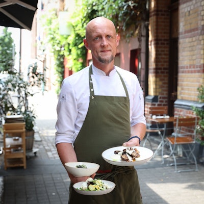 Mann in Kochkleidung steht vor einem Restaurant und hält drei Teller in der Hand