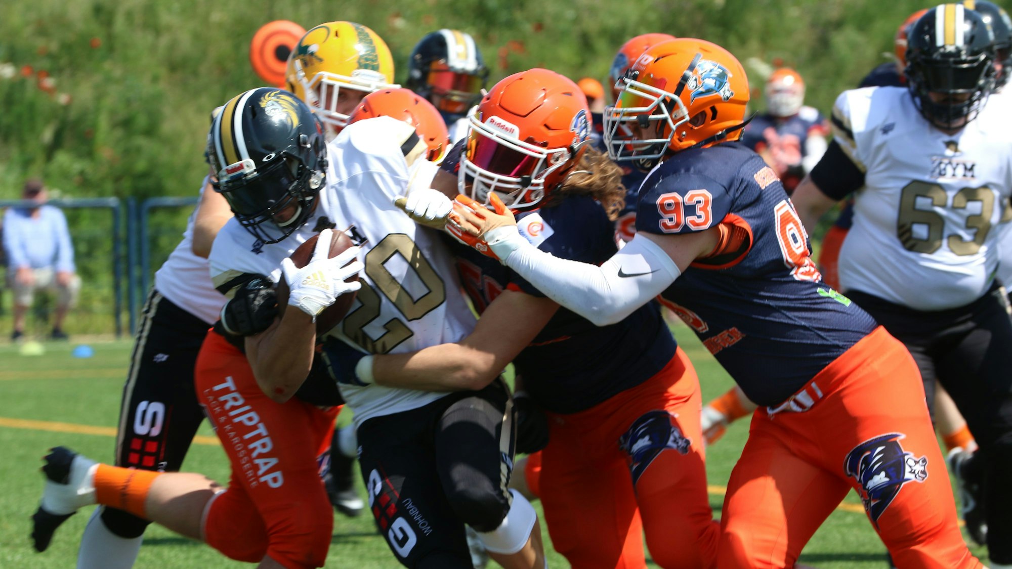 Das Bild zeigt eine Football-Spielszene: Ein Spieler im weißen Trikot hat den Ball und wird von mehreren gegnerischen Spielern bedrängt.