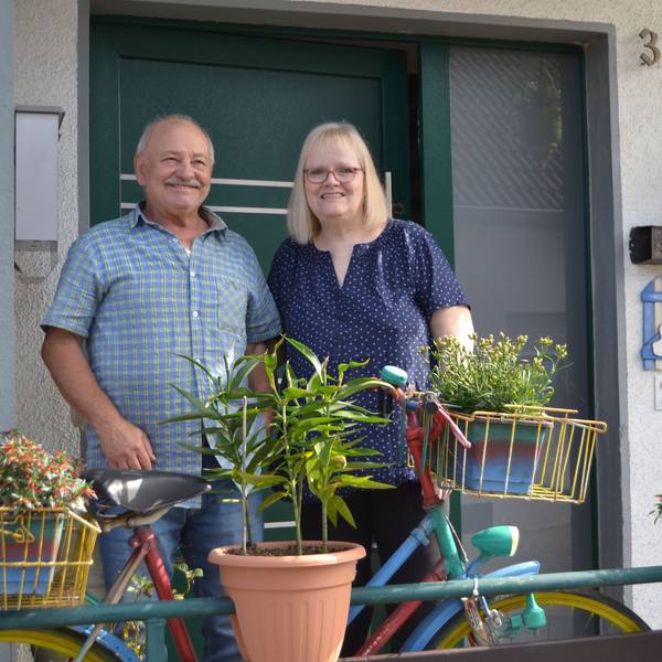 Ein Ehepaar steht vor einer Haustüre. Vor ihnen steht ein bepflanztes Fahrrad.