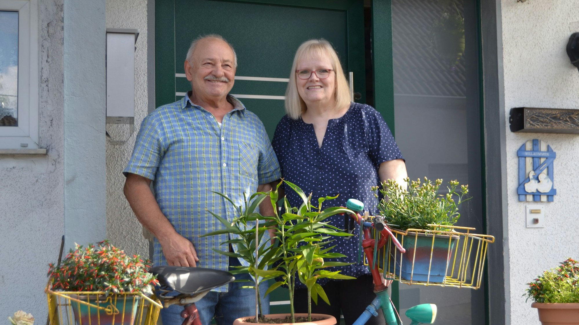 Ein Ehepaar steht vor einer Haustüre. Vor ihnen steht ein bepflanztes Fahrrad.
