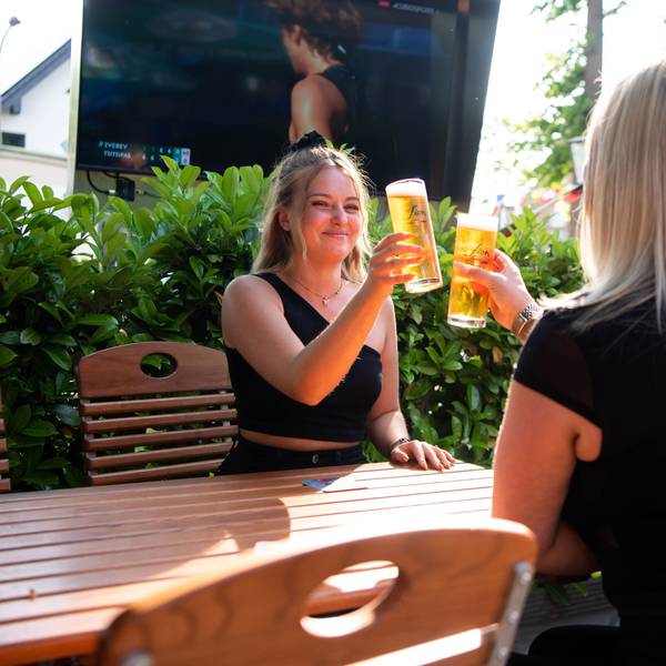 Zwei Frauen sitzen an einem Biergartentisch und stoßen mit einem Bier an.