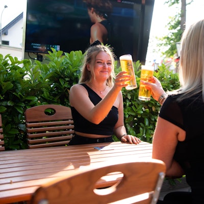 Zwei Frauen sitzen an einem Biergartentisch und stoßen mit einem Bier an.