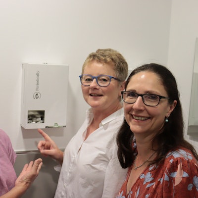 Drei Frauen präsentieren einen Spender für kostenlose Menstruationsartikel.