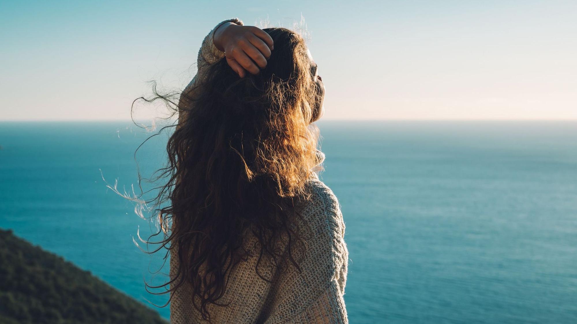 Eine Frau steht am Meer und streckt den Kopf Richtung Sonne.
