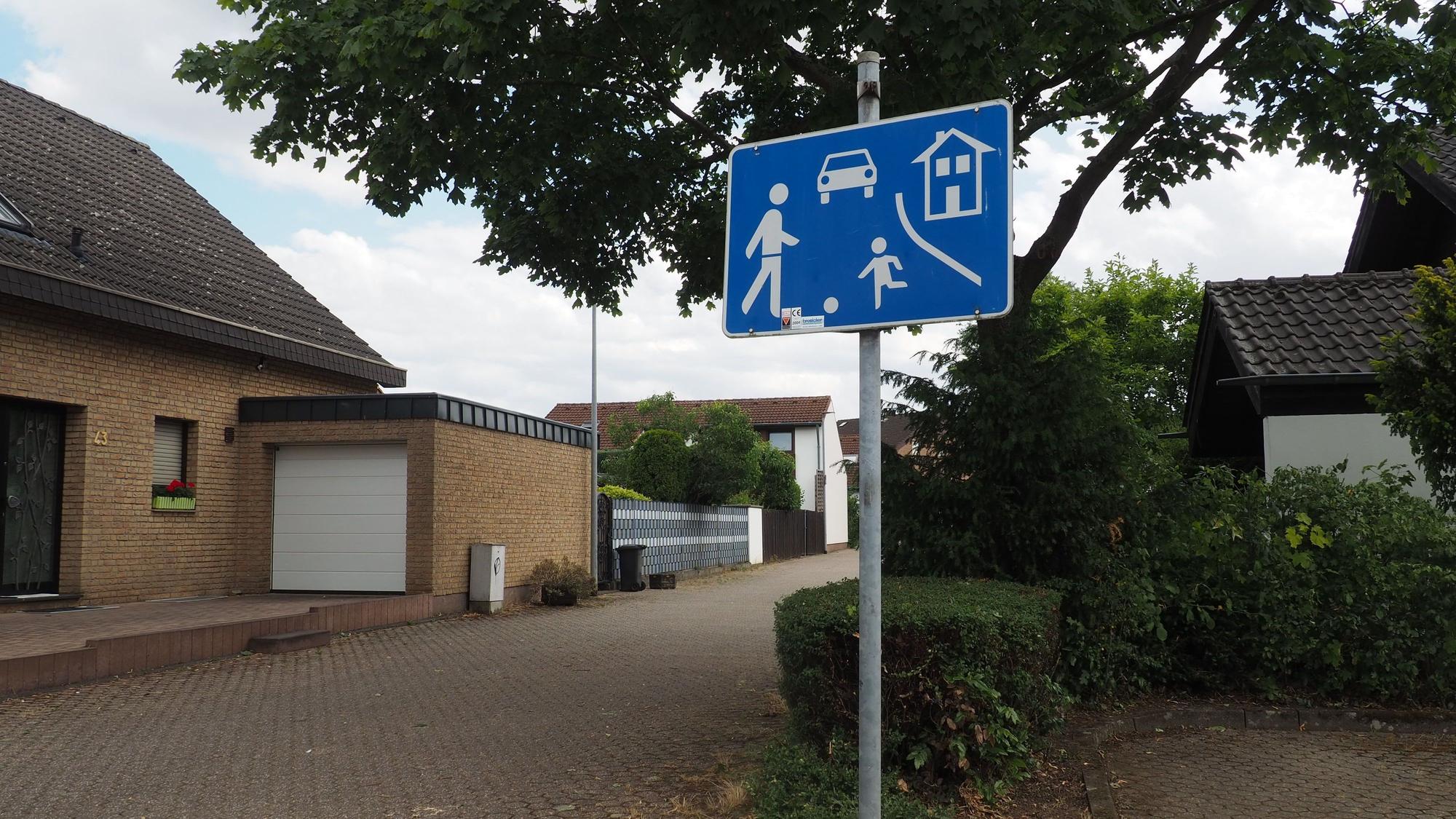 Ein blaues Verkehrsschild steht an einer Einmündung zu einer Straße in einem Wohngebiet.