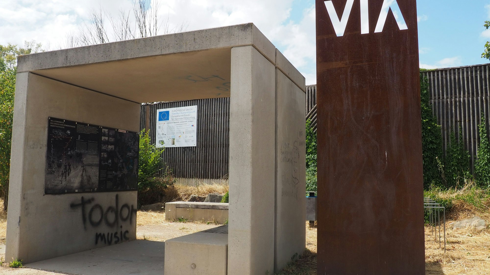 Ein offenes Bauwerk aus Beton spendet Schatten, daneben steht eine Stele aus verrostetem Metall.