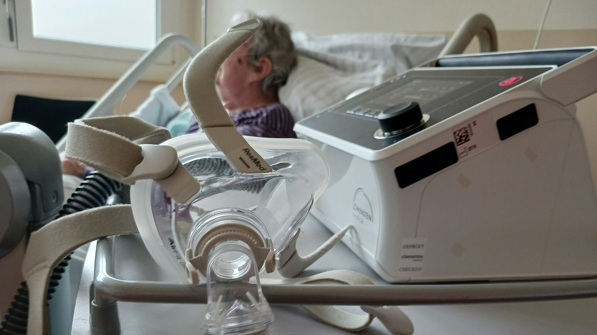 Eine Seniorin liegt in einem Bett im Krankenhaus. Vor ihr steht ein Atemgerät.