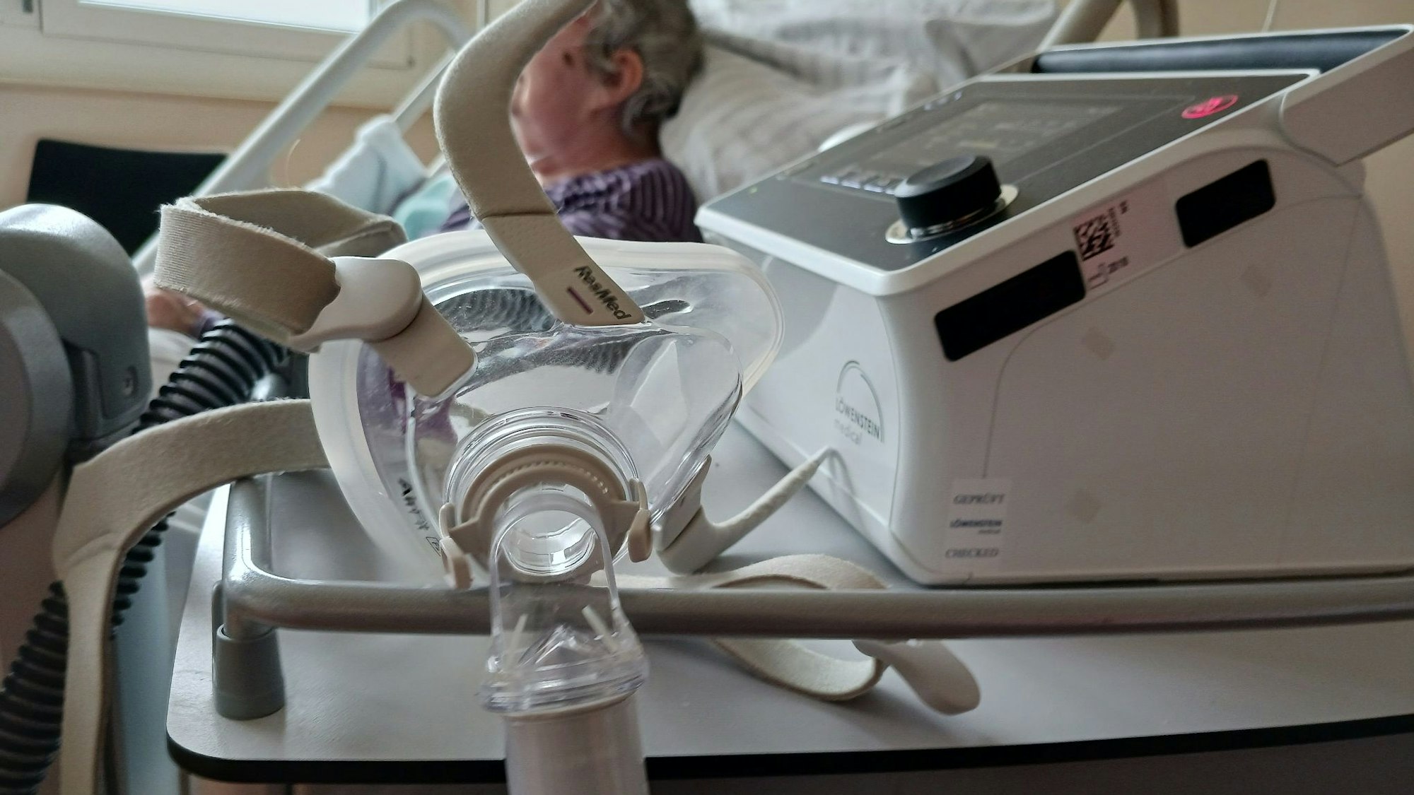 Eine Seniorin liegt in einem Krankenhausbett, im Vordergrund hängt eine Atemmaske am Beatmungsgerät. (Symbolfoto)
