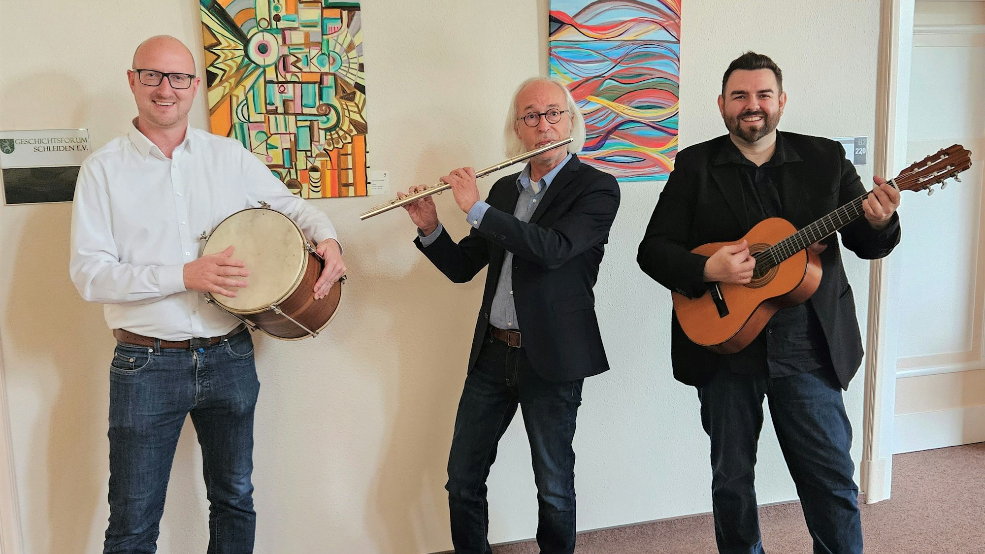 Das Foto zeigt Bernhard Vorhagen mit einer Querflöte, Ingo Pfennings mit einer kleinen Trommel und Peter Rack mit einer Gitarre. Sie stehen vor einer Wand, an der zwei bunte Bilder hängen.